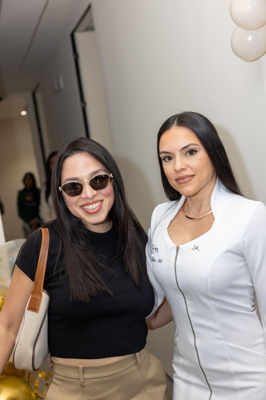 Two people stand smiling side-by-side in an indoor setting; one wears a black top and sunglasses, the other a white coat.