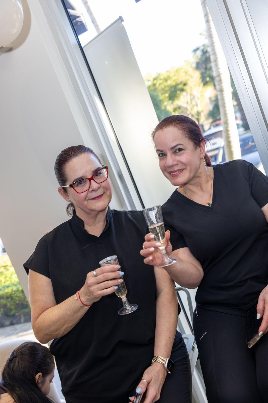 Two people in black tops hold up champagne flutes for a toast, smiling in a brightly lit indoor setting.