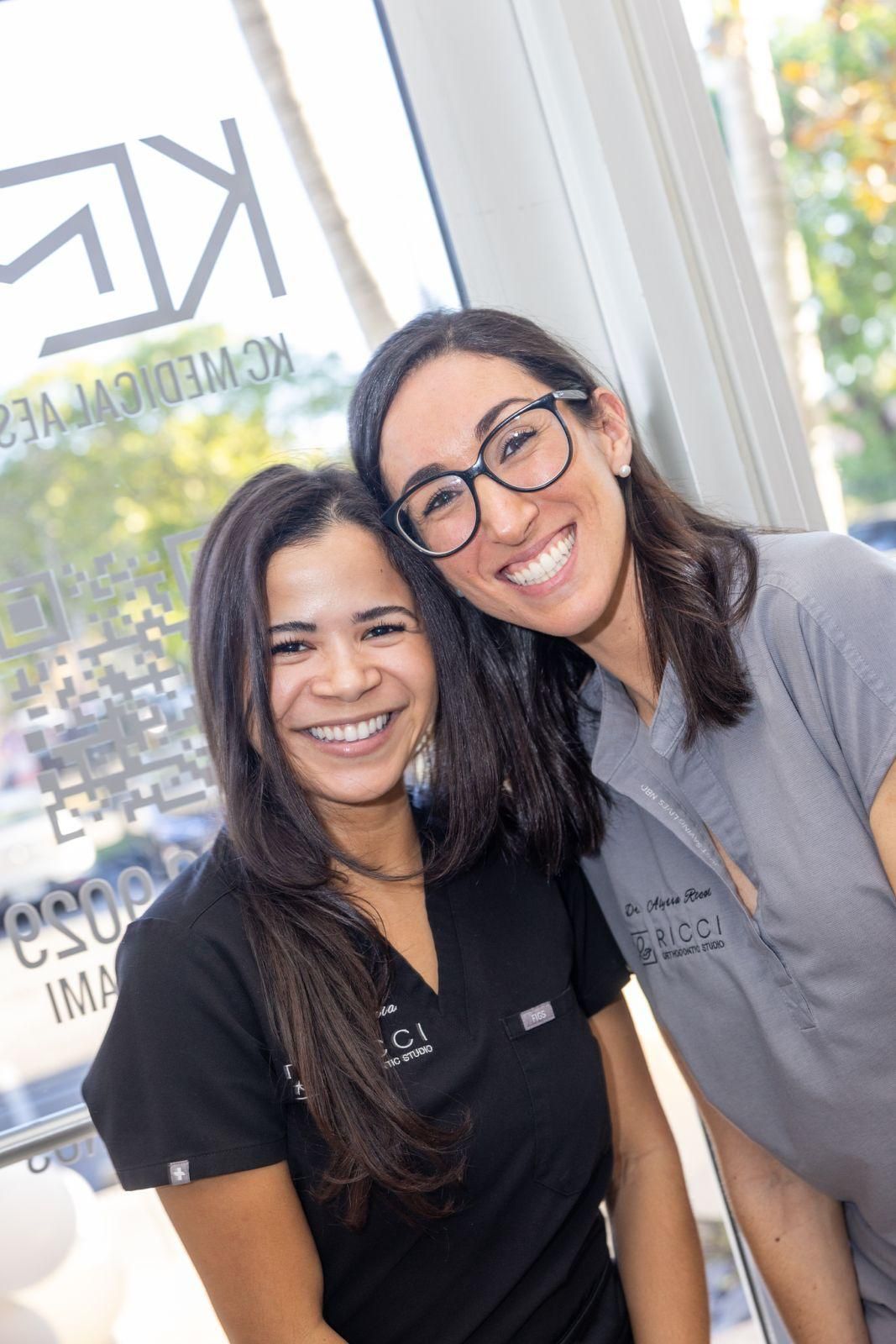Two smiling professionals in scrubs stand inside an office near a glass door with a medical logo in the background.