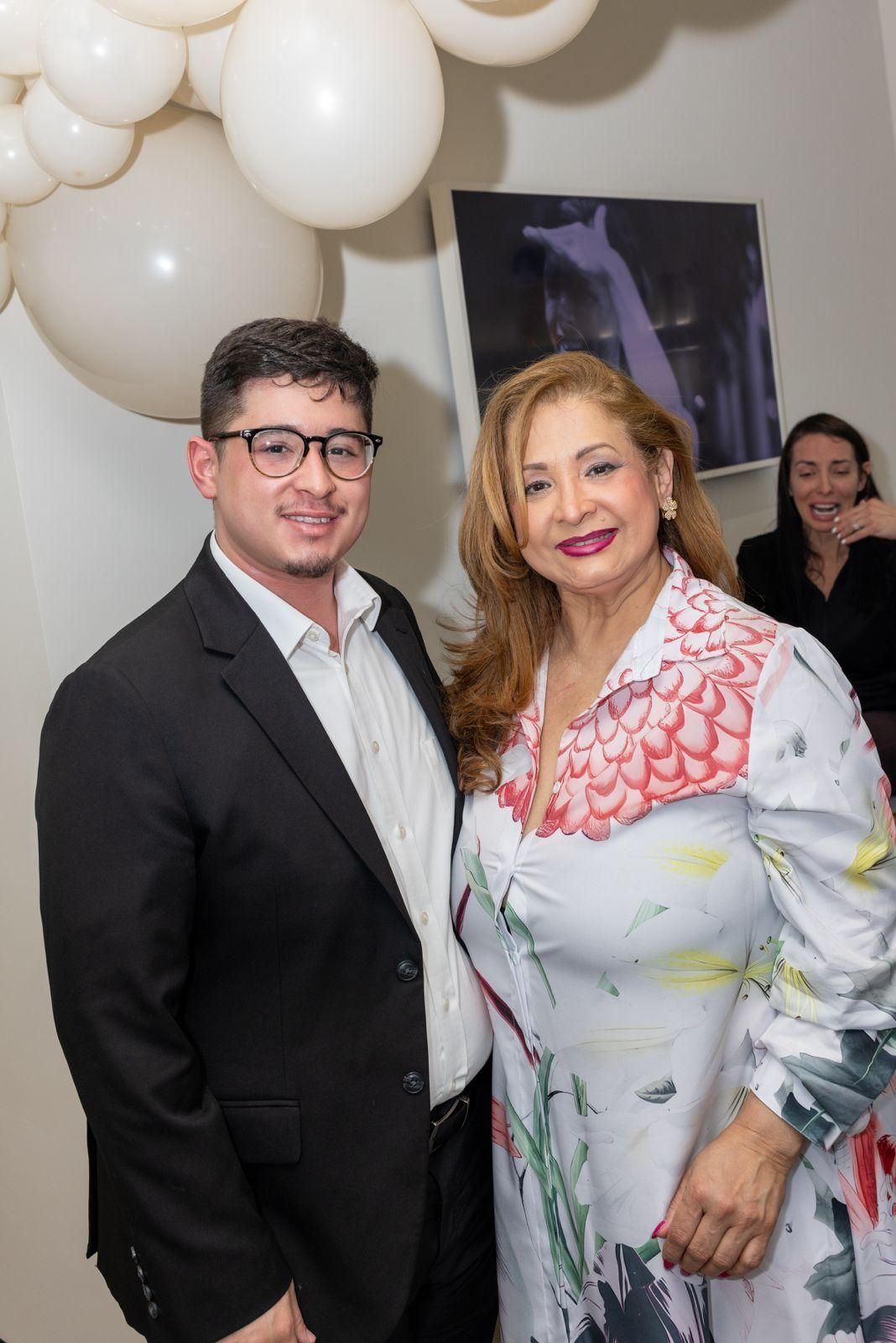 A smiling person in a suit stands next to a woman in a patterned dress, with balloons in the background.