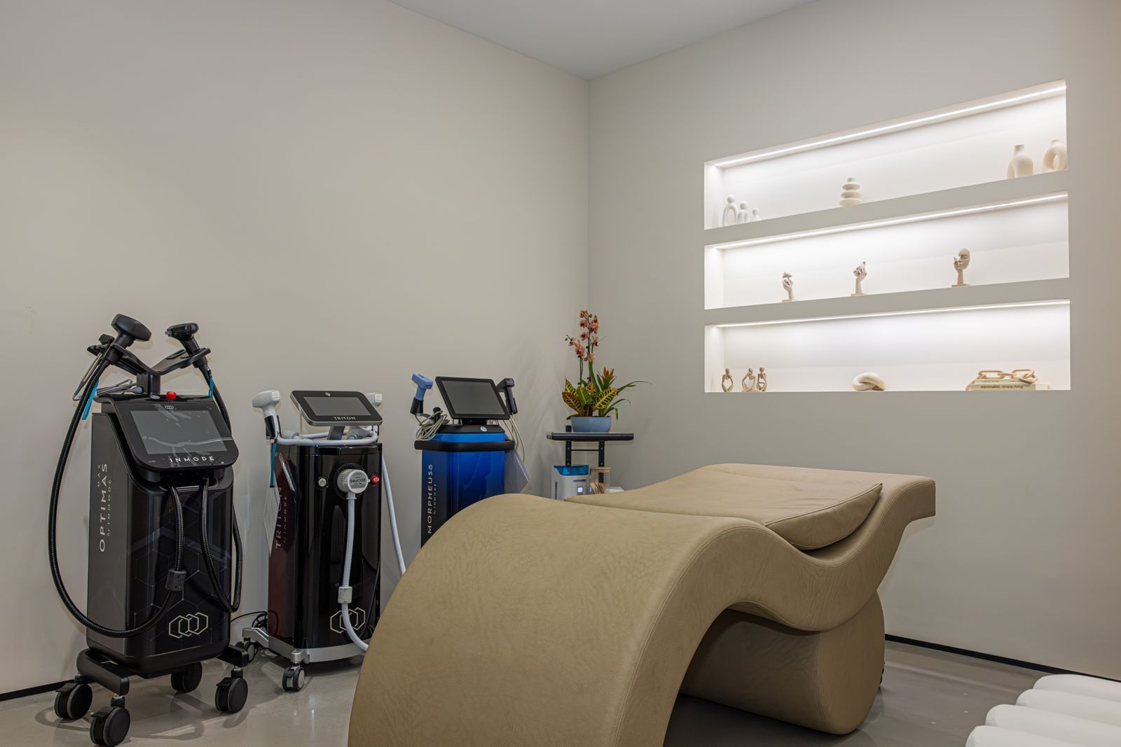 A treatment room with three medical aesthetic devices on the left, a curved beige lounge chair, and illuminated shelving.