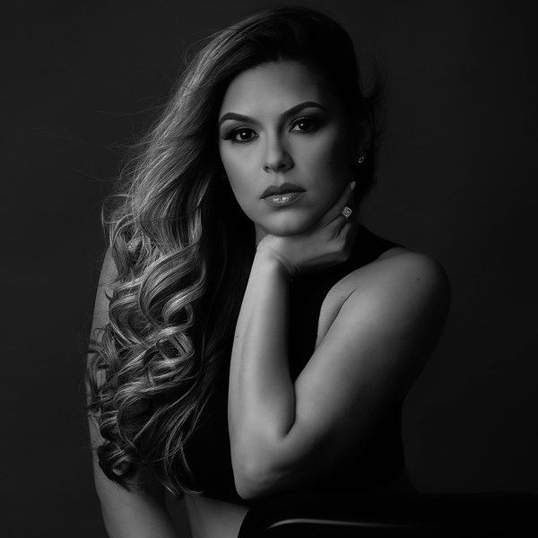 Black and white portrait of a person with long wavy hair, resting their chin on their hand, against a dark background.