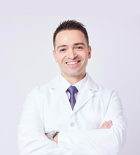 A professional in a white lab coat and tie smiling with arms crossed against a light background.