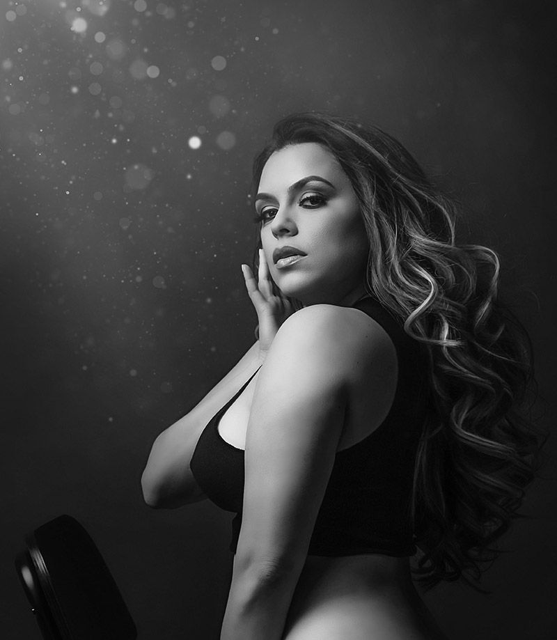 A person with long wavy hair, wearing a black top, poses against a dark, bokeh-lit background.