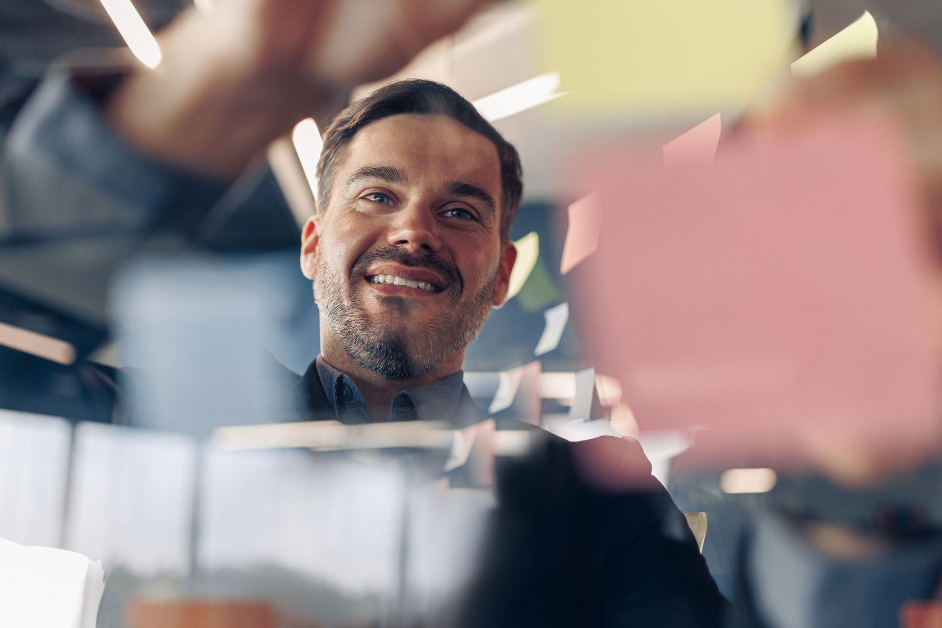 Man smiles, placing sticky notes on a clear surface, likely brainstorming in an office setting.
