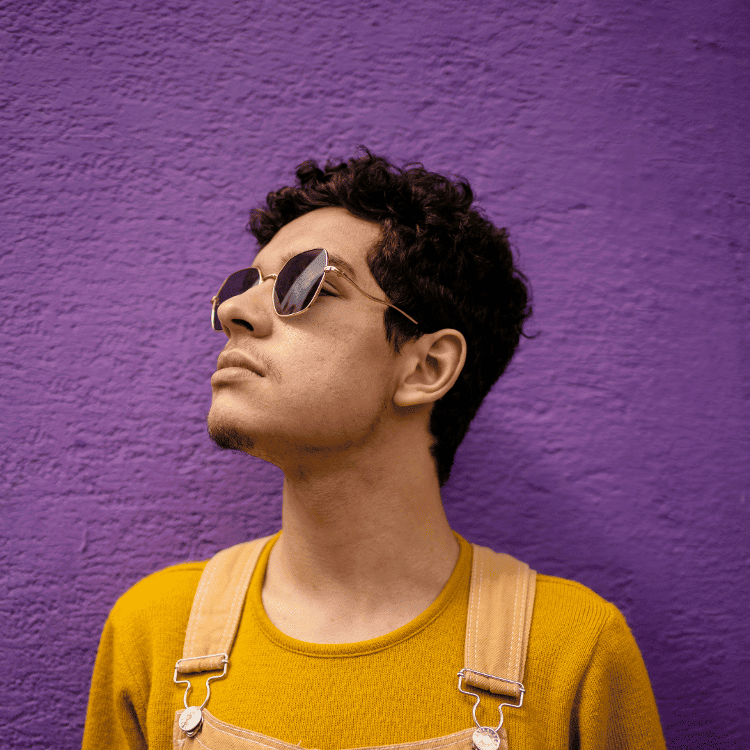 AI Engineer in sunglasses and mustard sweater against a purple wall, looking upward.