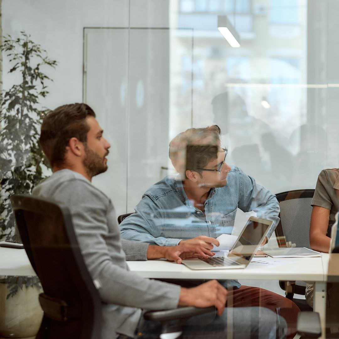 People in a glass-walled meeting room discussing around a table with a laptop and documents