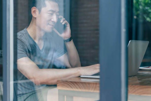 Man in grey shirt on phone, working on laptop at a cafe table, seen through window.