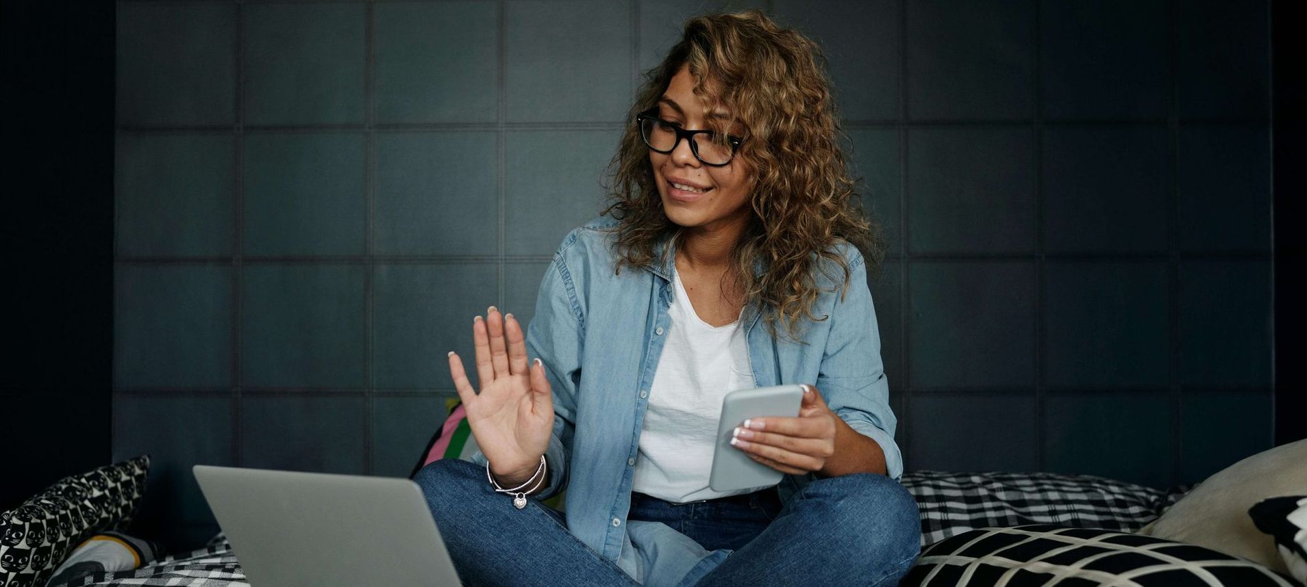 Woman with glasses waves at a screen while holding a tablet. She sits on a bed with a laptop.