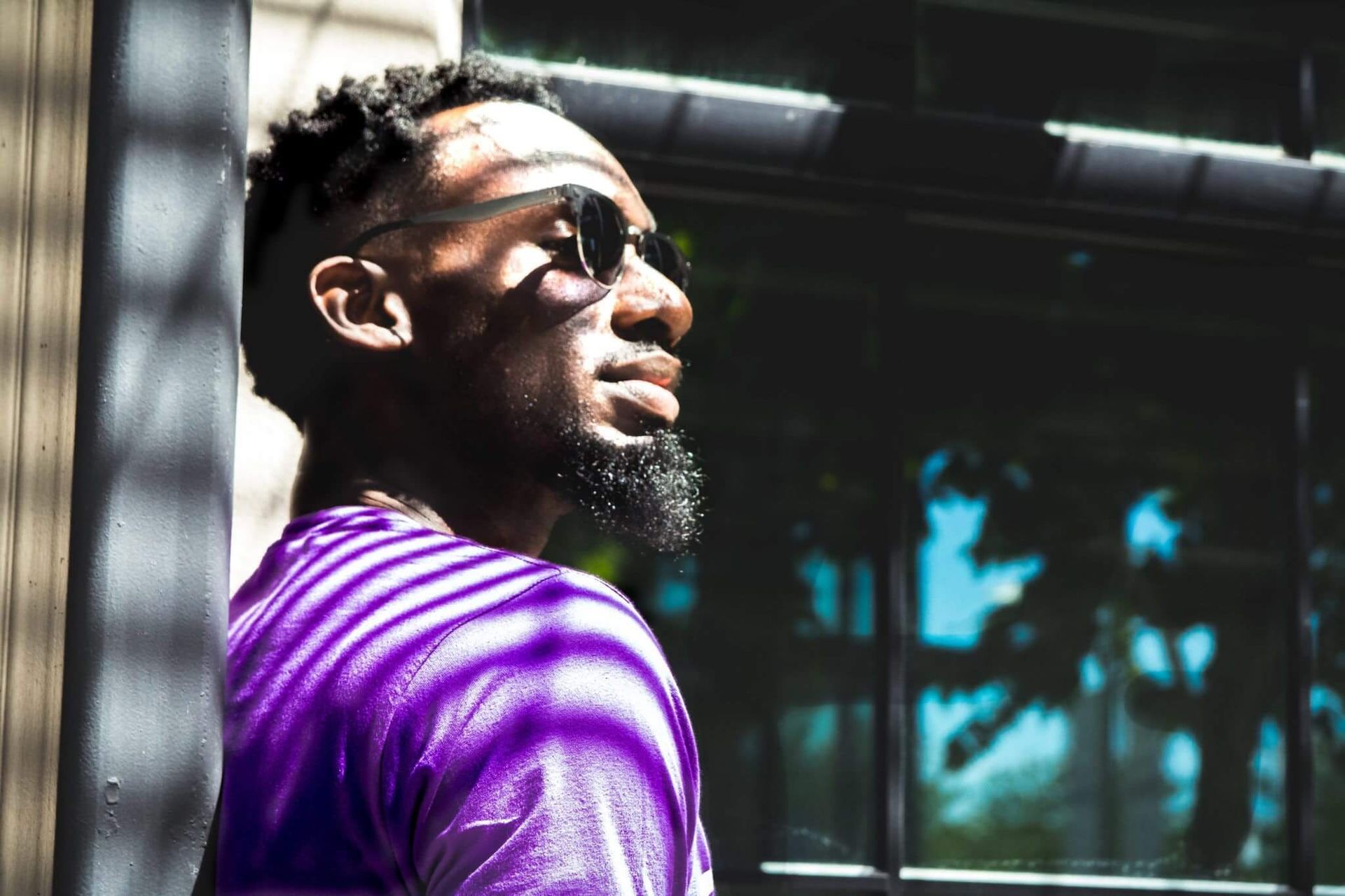 Man in sunglasses, purple shirt, looking up. Shadow stripes, window background, sunny.