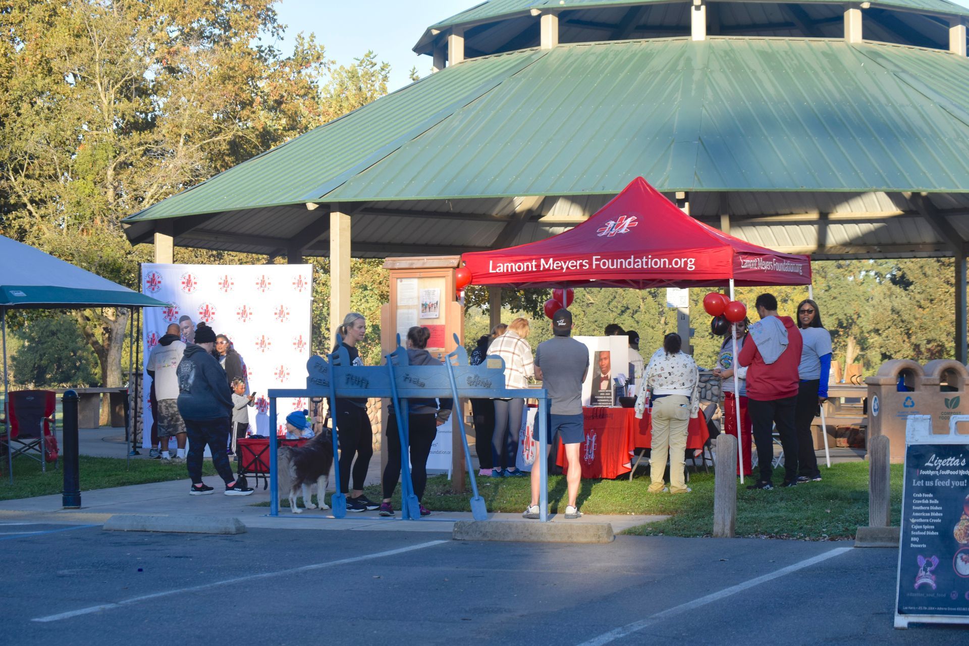 People gather at an outdoor event with red tent and gazebo. A dog on a leash, tables, trees.