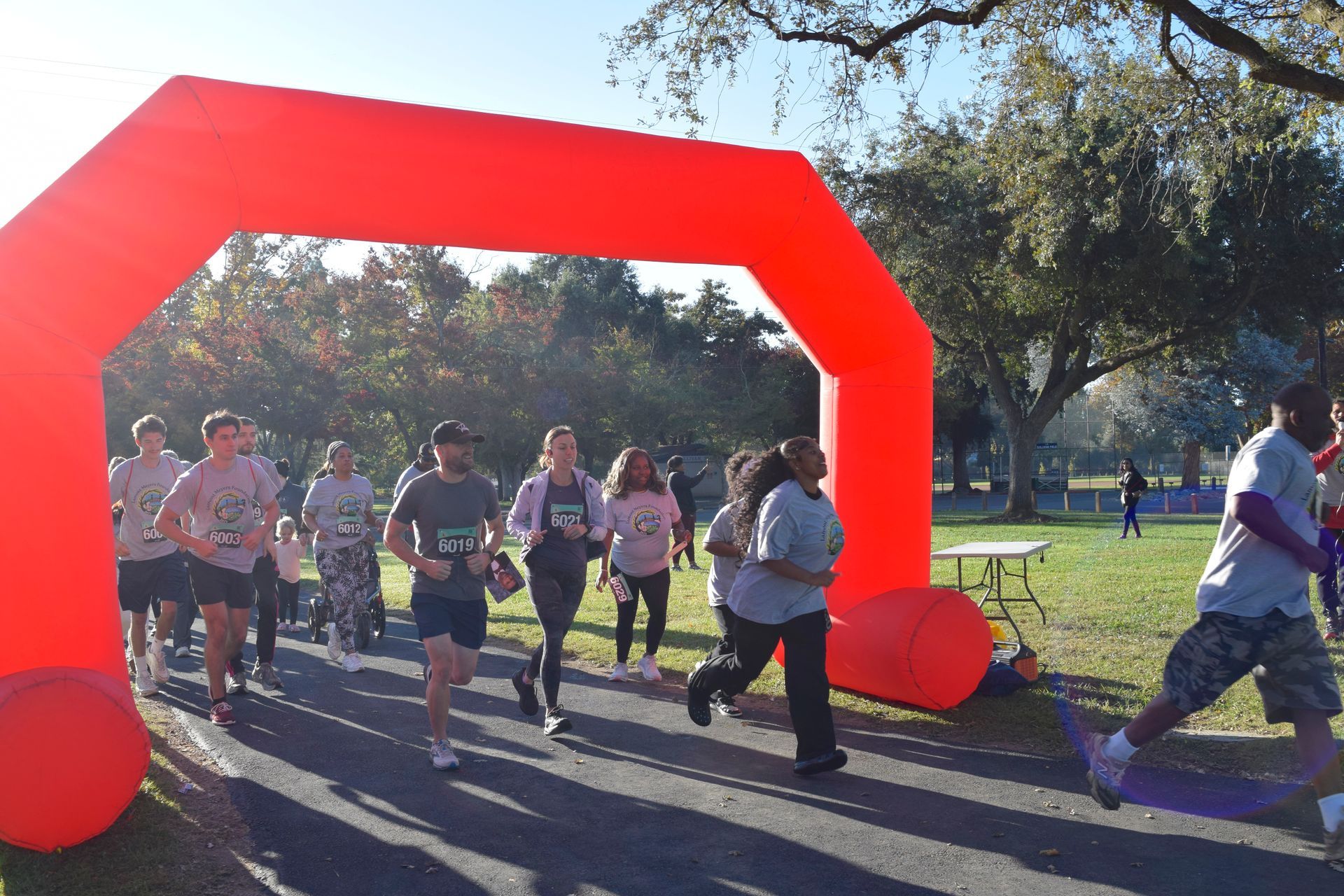 Runners start a race under a bright red archway in a park on a sunny day.