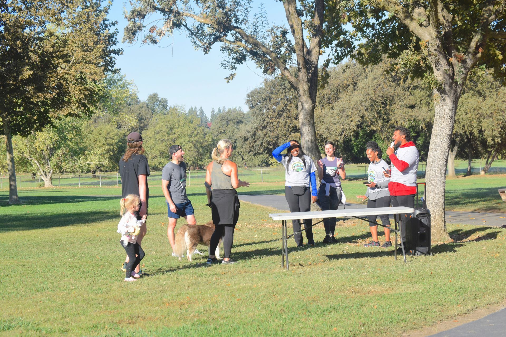 People gather near a table under trees in a park. Some hold papers; a dog is present.