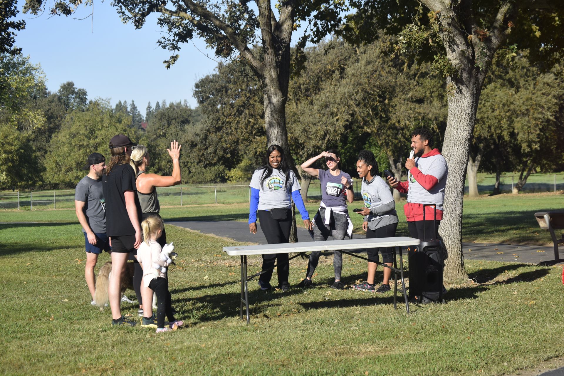 Group of people at an outdoor event, some at a table, under trees. One person has a microphone.