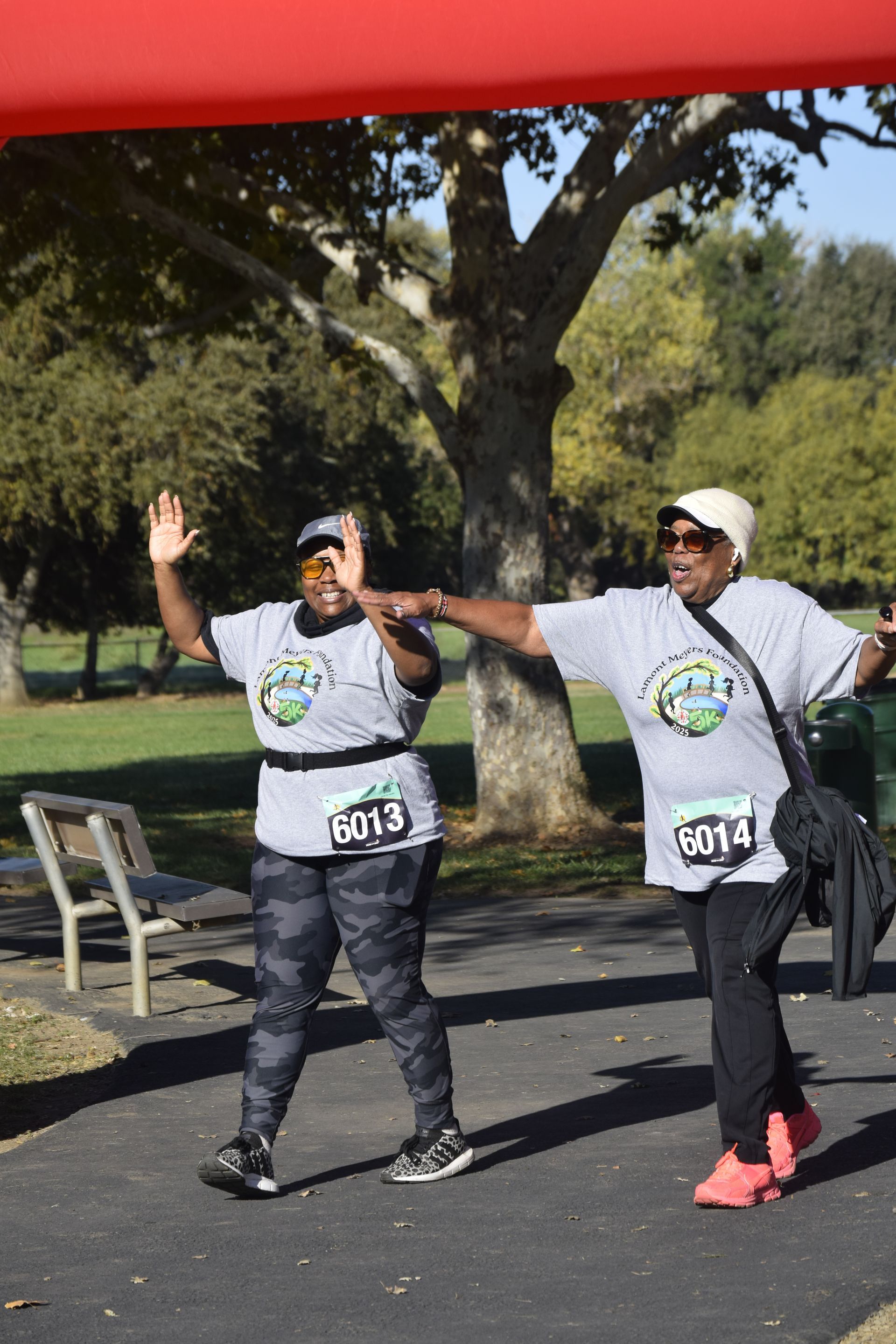 Two women in matching shirts finish a race, raising arms under a red banner; outdoors on a path, sunny day.