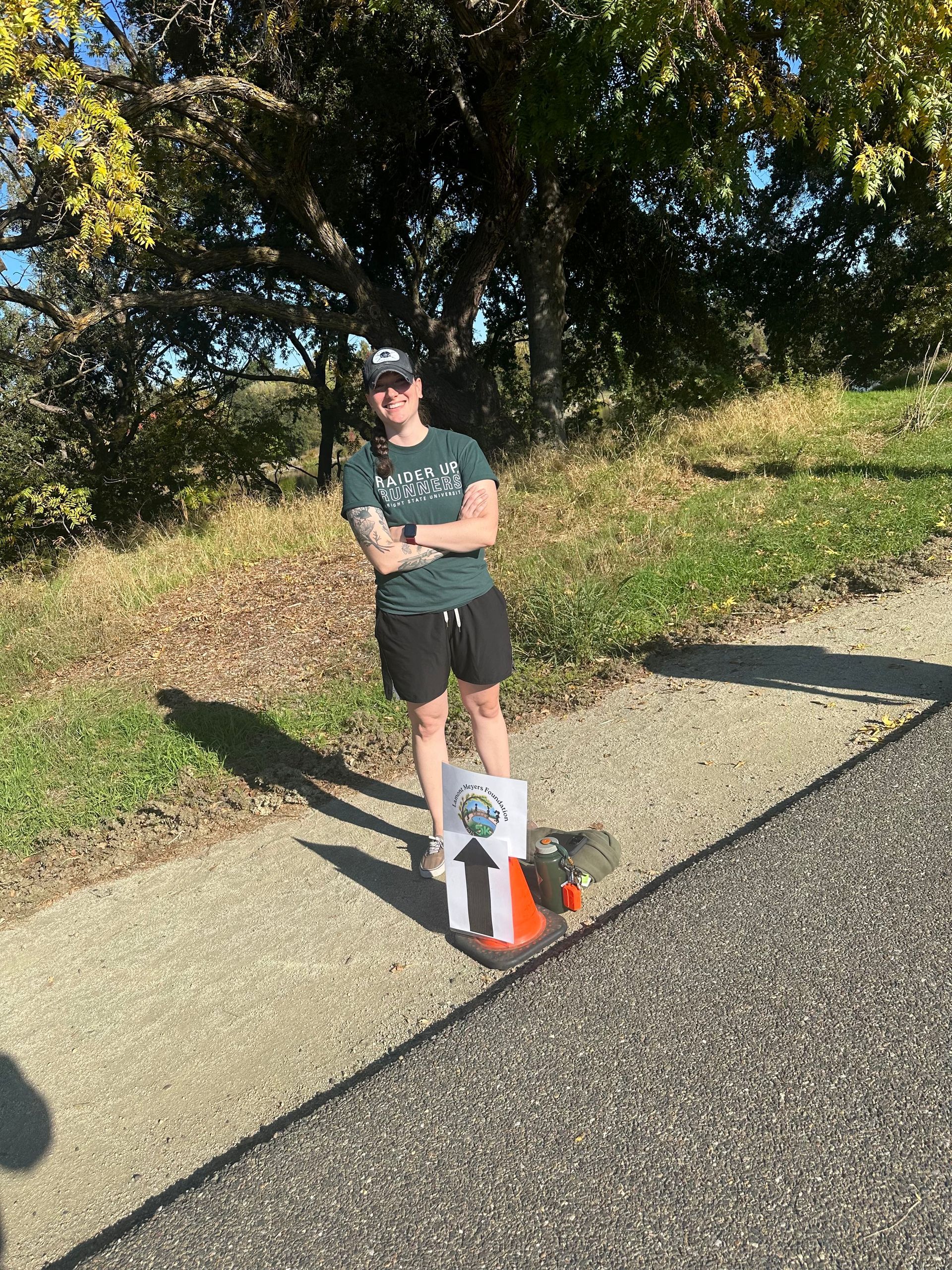 Person standing next to a sign with an arrow on a sidewalk. They are outside with a tree behind them.