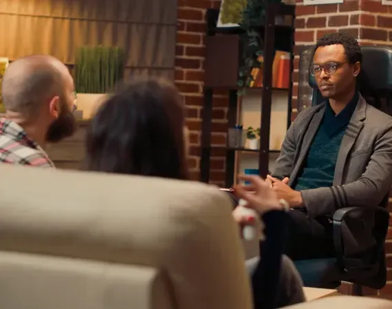 Couple in therapy session with therapist. Brick wall and bookshelves in background.