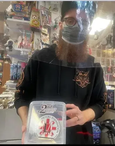 Man with a beard, mask, and glasses holding a clear plastic box with a logo. Inside a store, counter visible.
