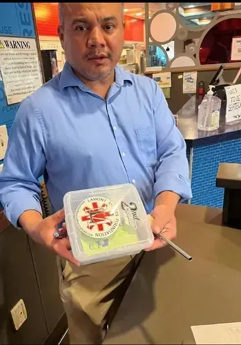 Man holding a clear plastic container with a sticker, next to a cash register.