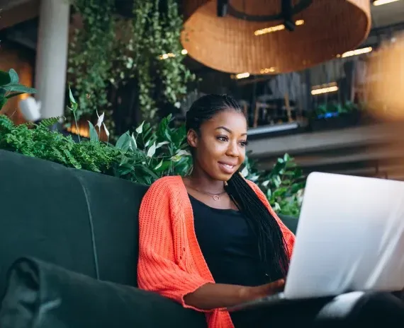 Woman using a laptop, sitting on a dark green couch, wearing orange sweater in a modern setting with greenery.