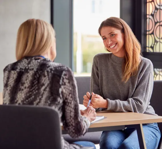 Woman smiling, seated, talking to another woman at a table indoors.