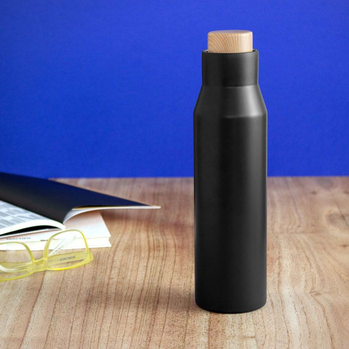 Black water bottle with wooden cap on a desk beside an open book and yellow glasses