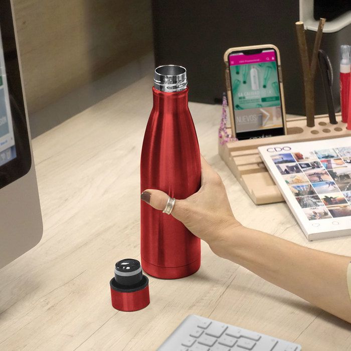 Hand reaching toward a red insulated bottle on a desk beside a keyboard and phone display