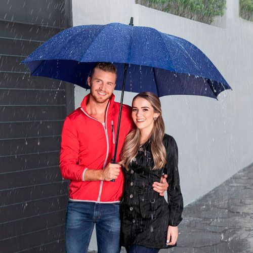 Two people under a blue umbrella in the rain, standing by a gray wall and smiling.