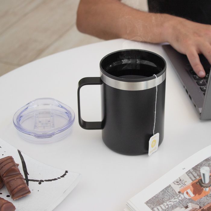 Black mug on a white table beside a clear lid, chocolate bar, and a person using a laptop