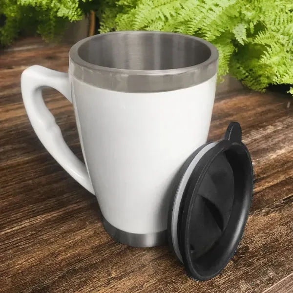 White insulated travel mug with stainless steel rim and black lid on a wooden table, fern in background