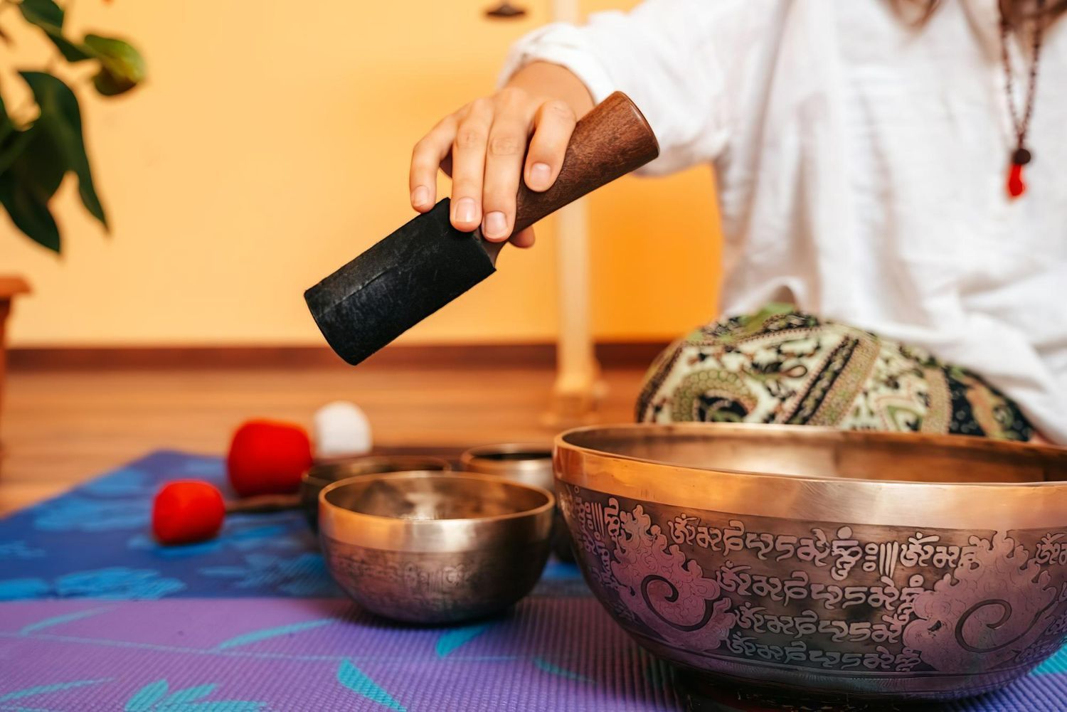 Person striking a singing bowl with a mallet beside other bowls on a colorful mat