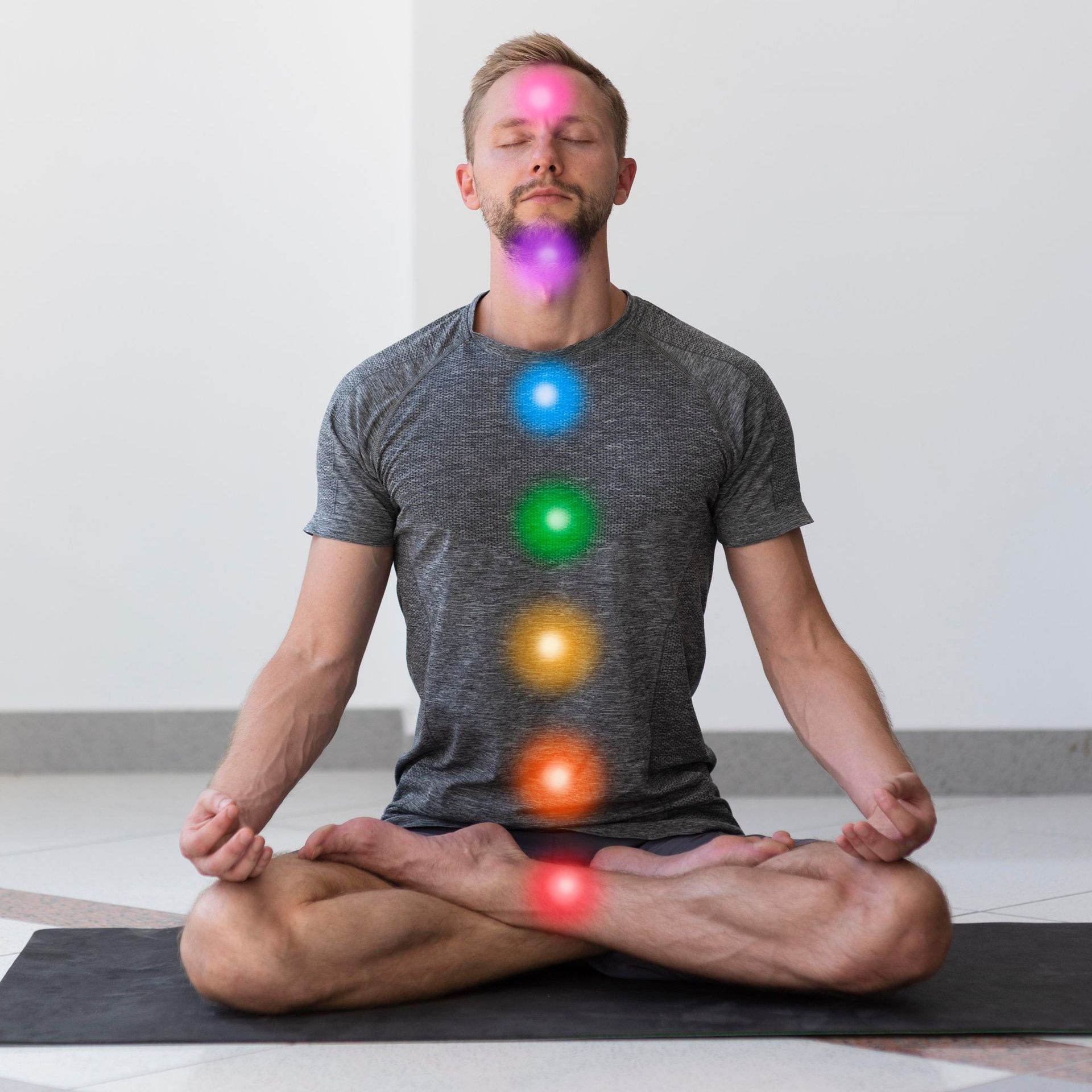 A man is sitting in a lotus position on a yoga mat with chakras glowing in the background.