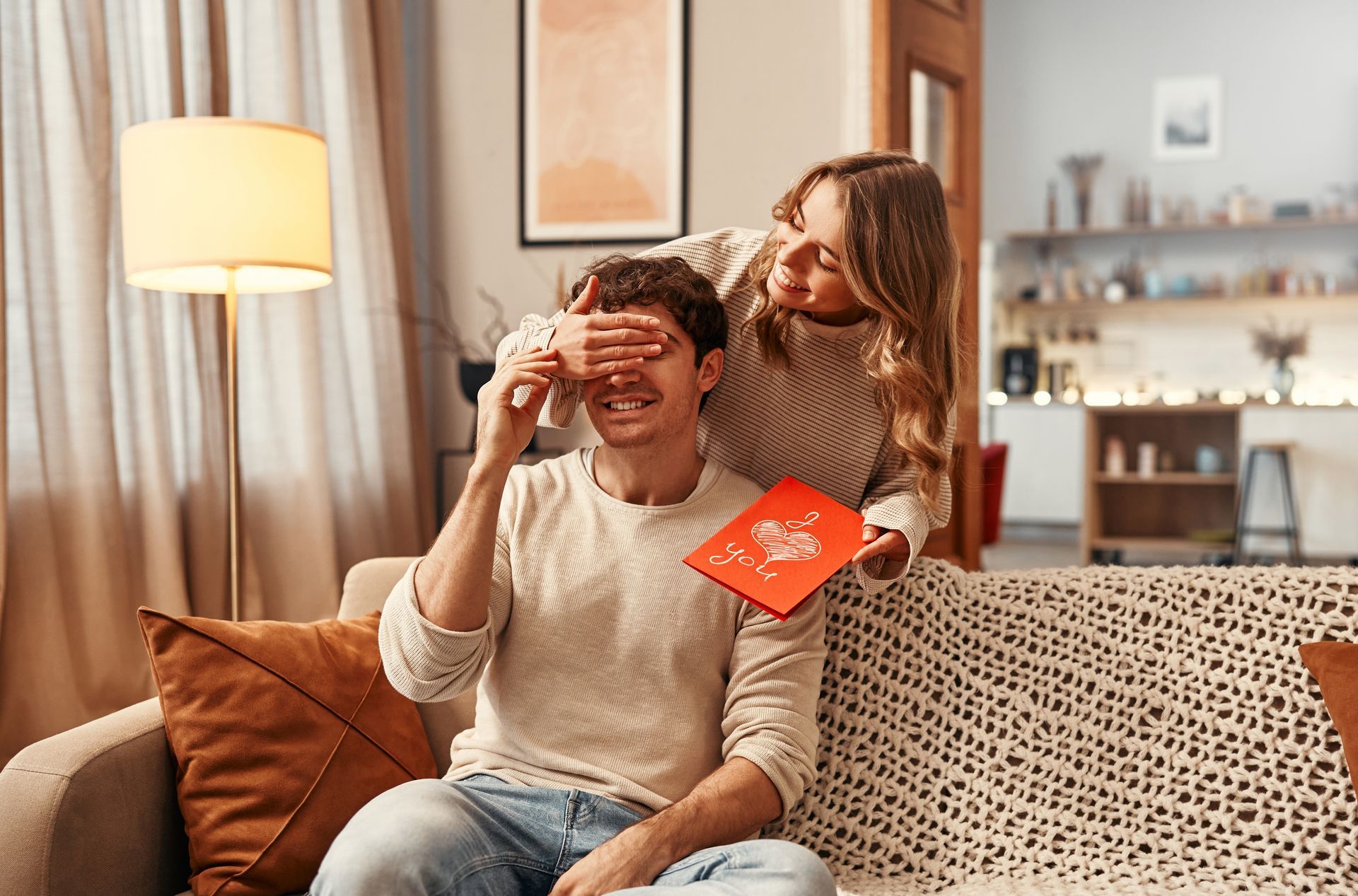 A woman is covering a man 's eyes while he is sitting on a couch.