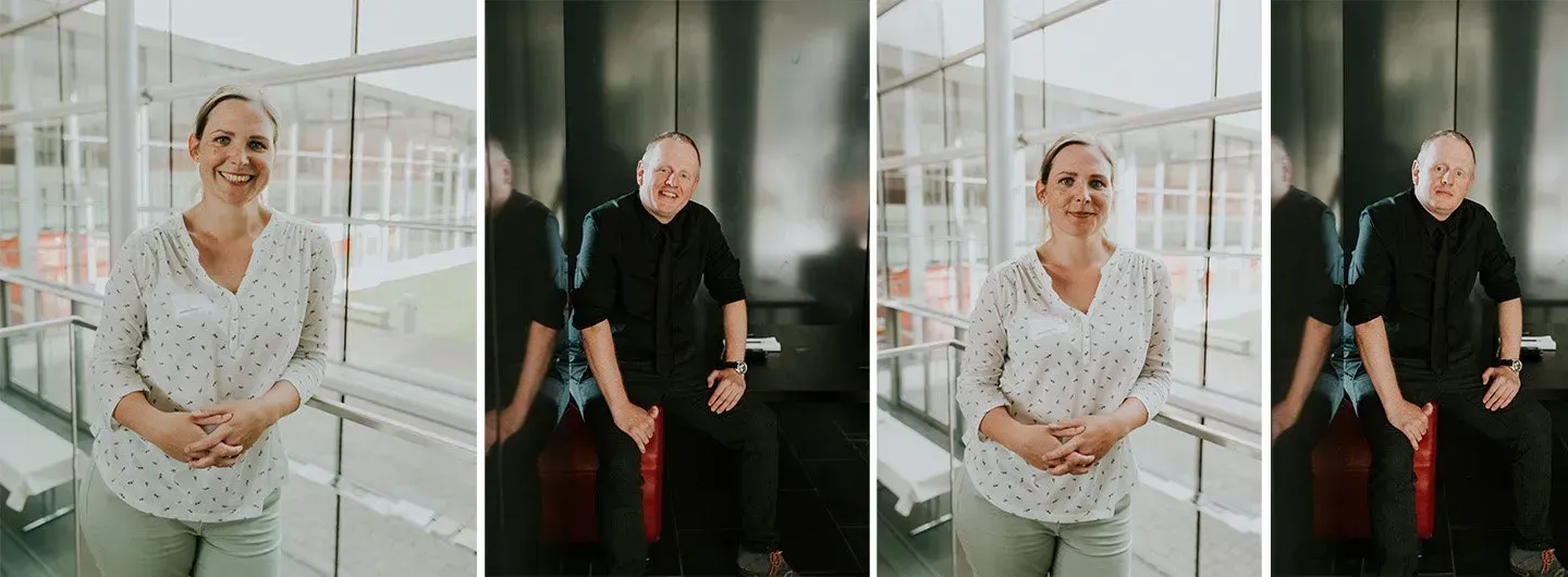 Eine Frau und ein Mann posieren in einem modernen Gebäude. Die Frau trägt ein gepunktetes Hemd und der Mann ein schwarzes Hemd.