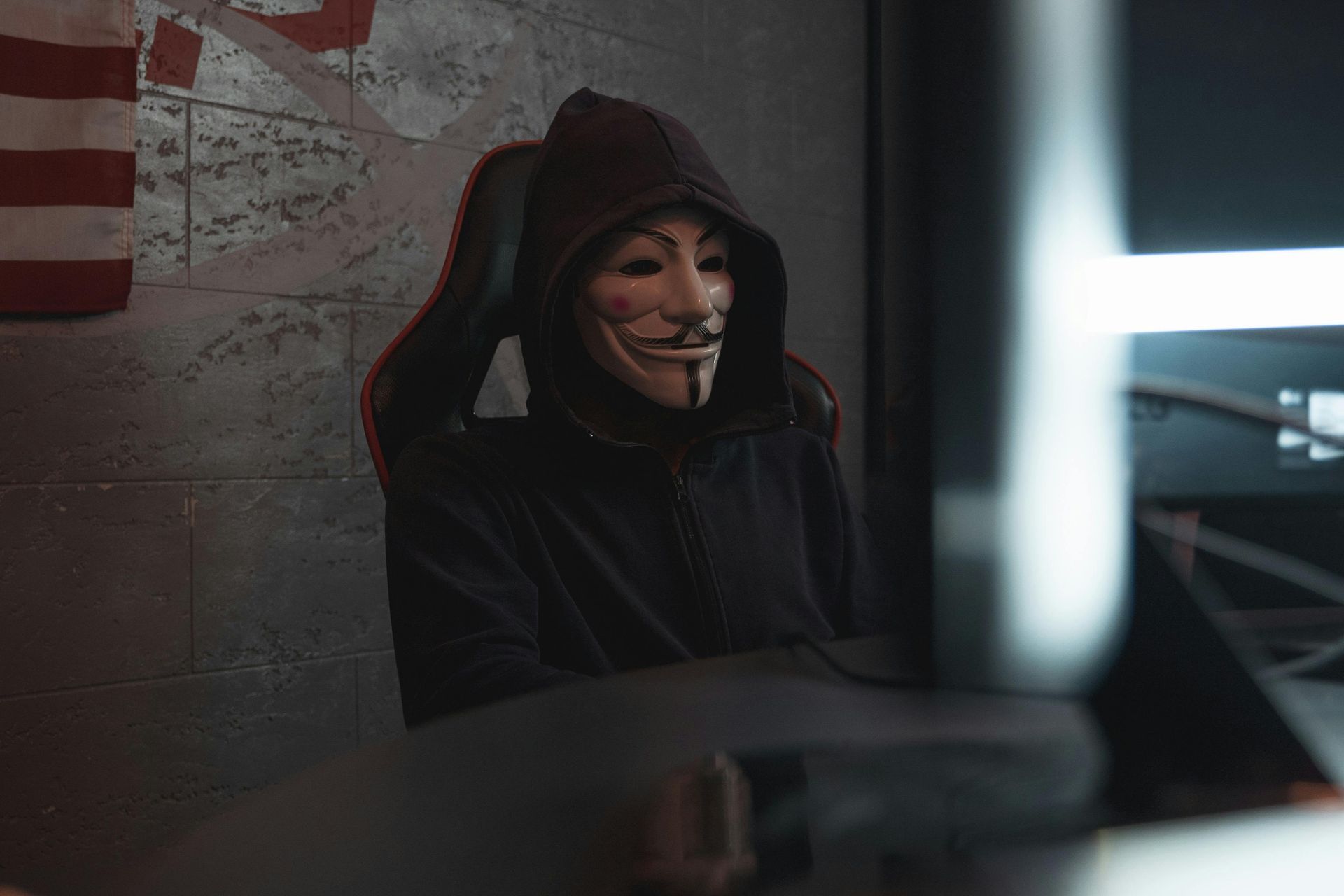 Person mit Guy-Fawkes-Maske und Kapuzenpullover, die in einem schwach beleuchteten Raum an einem Computer arbeitet, im Hintergrund die amerikanische Flagge.