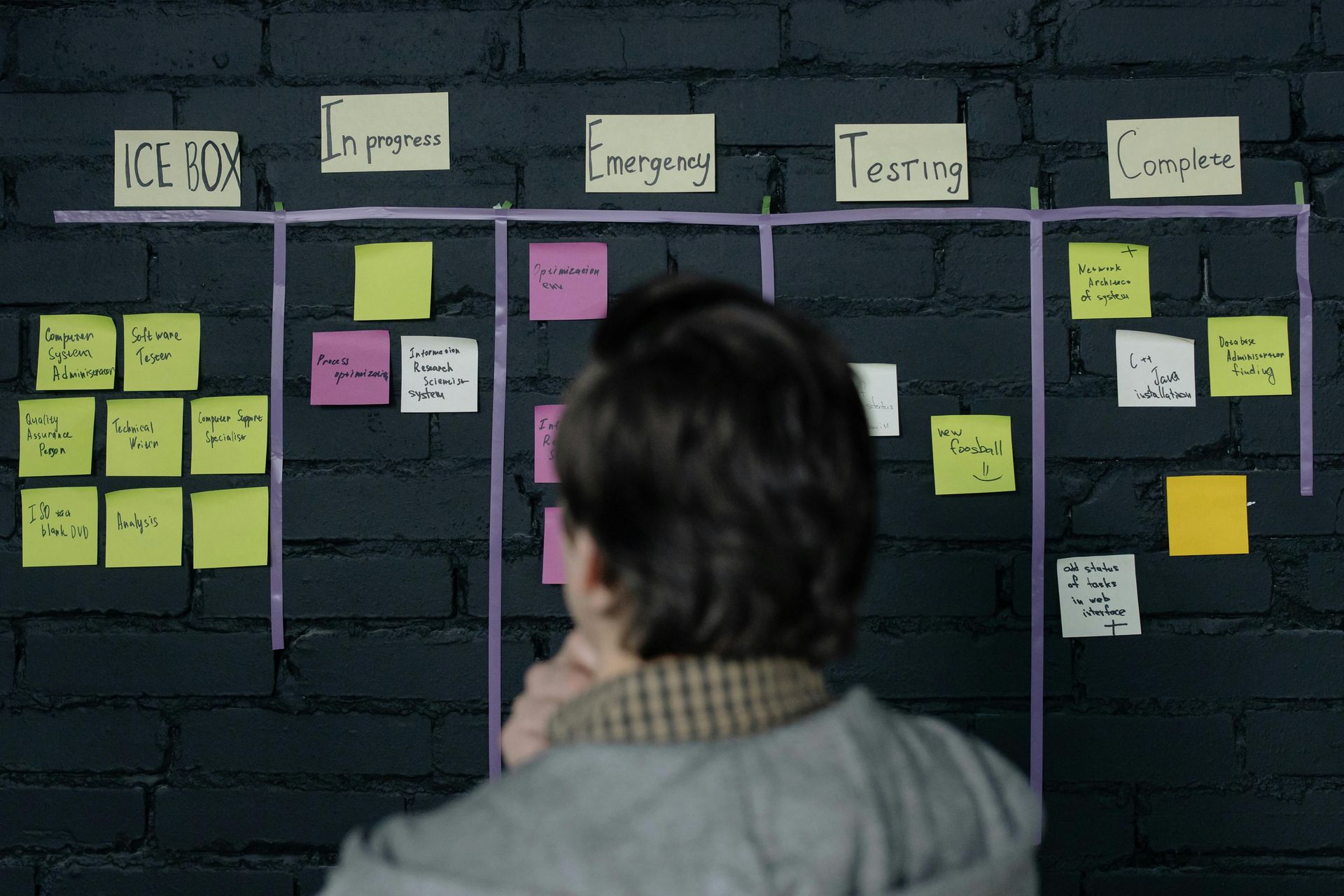Person betrachtet ein Kanban-Board mit Haftnotizen, nach Phasen geordnete Aufgaben auf einer dunklen Ziegelwand.