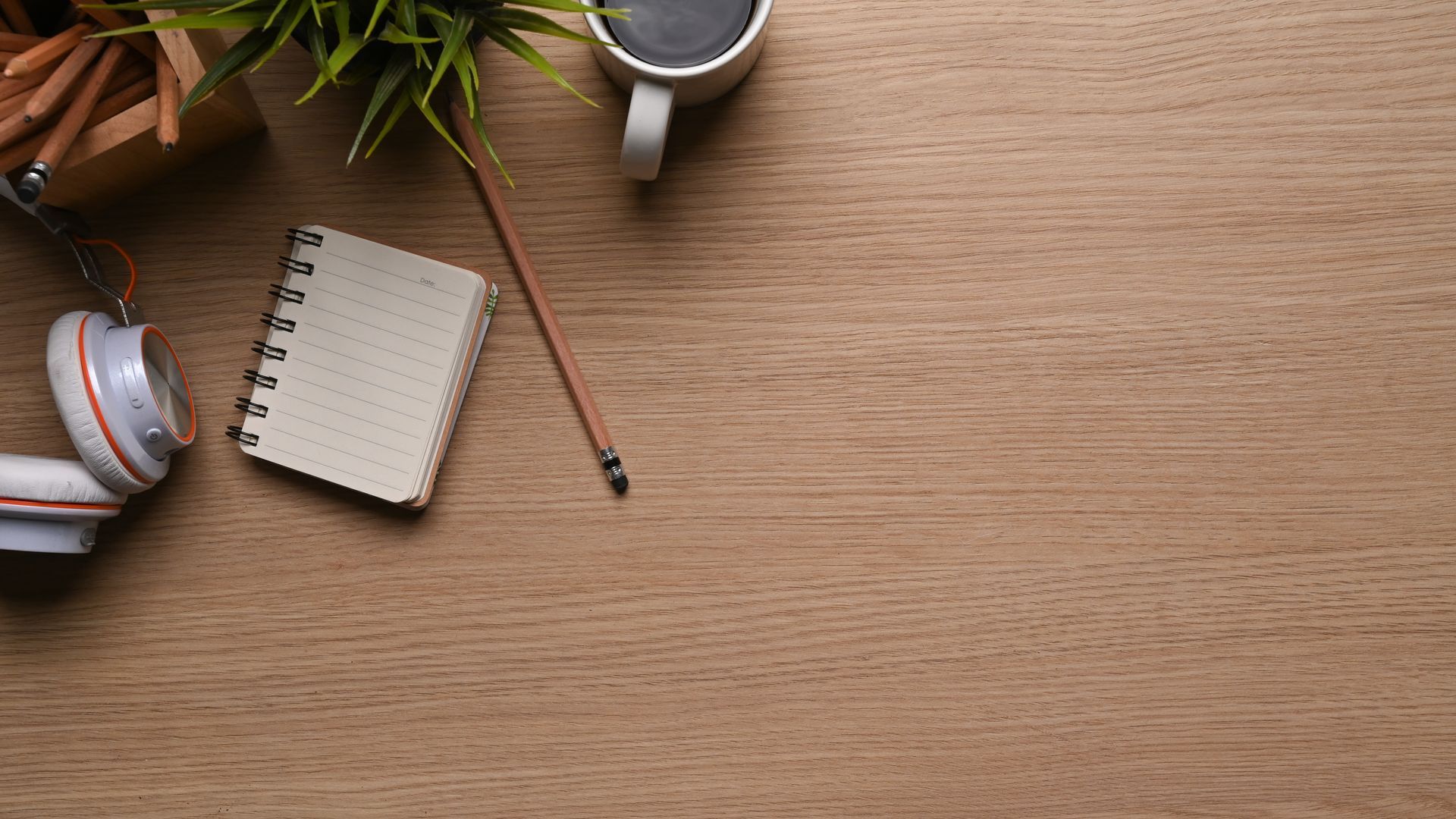 Wooden desk with notebook, pencils, headphones, coffee, and plant.