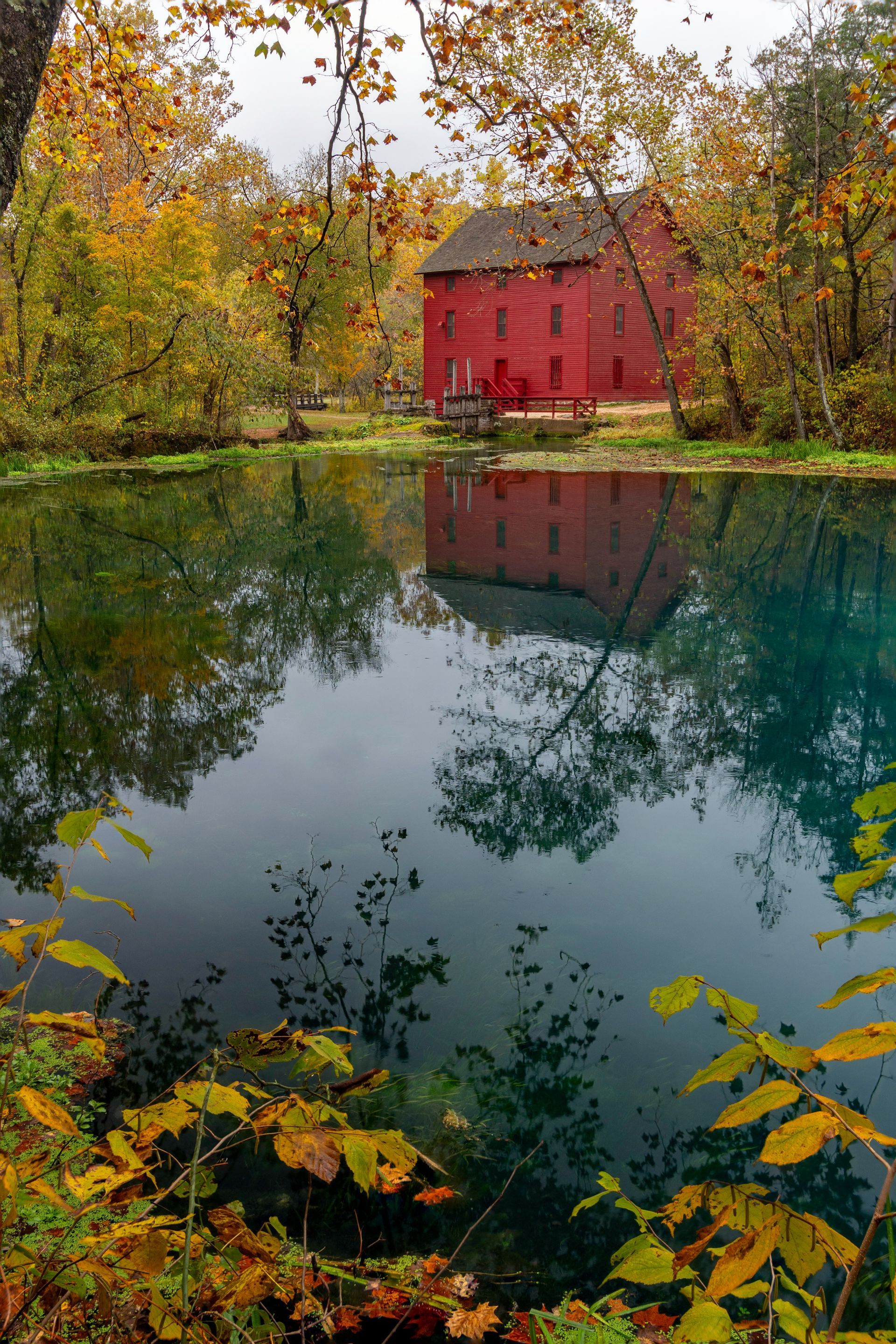 Red barn beside a calm pond, reflected in water, surrounded by colorful autumn trees.