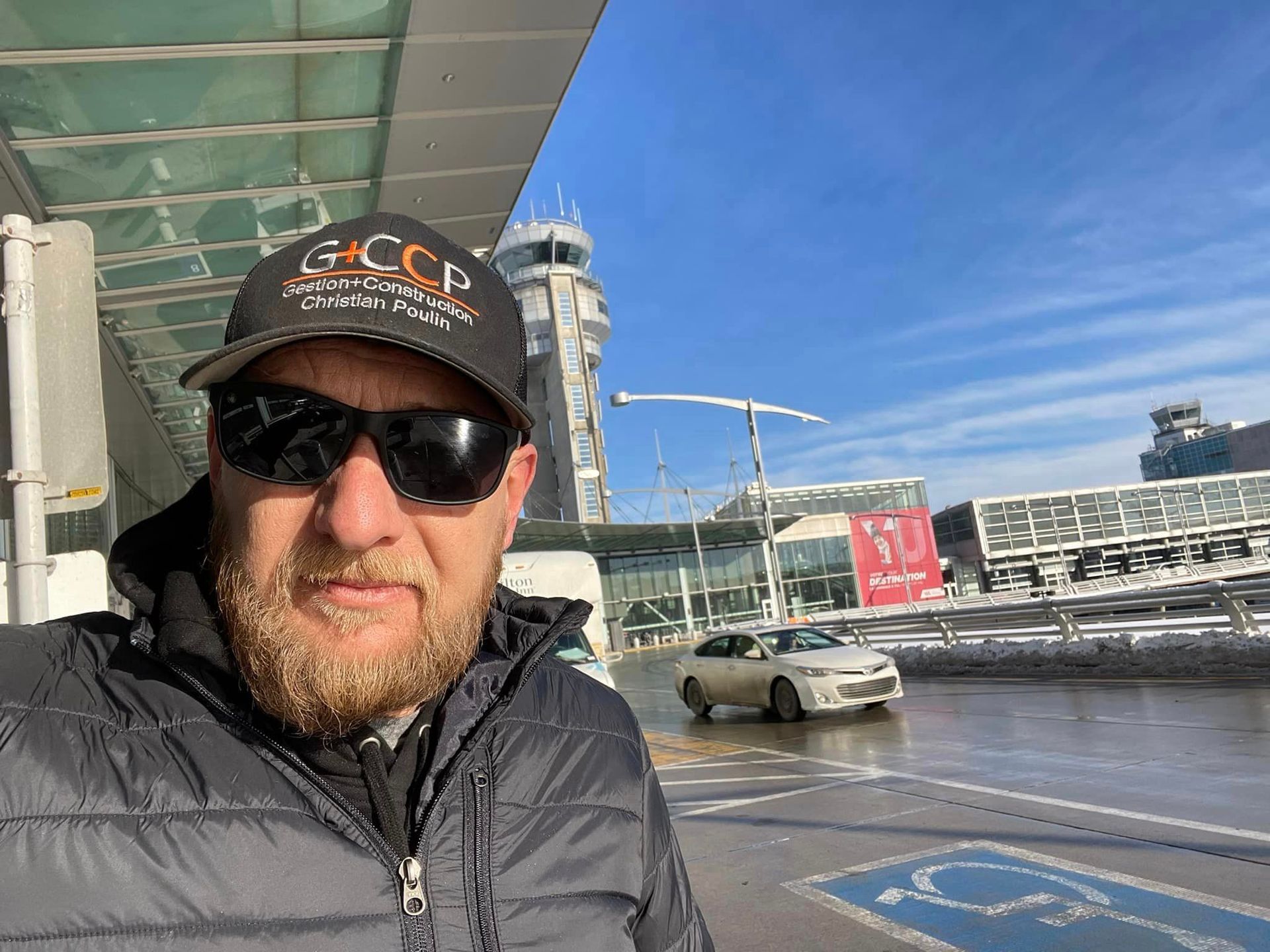Un homme portant un chapeau et des lunettes de soleil se tient devant un aéroport.