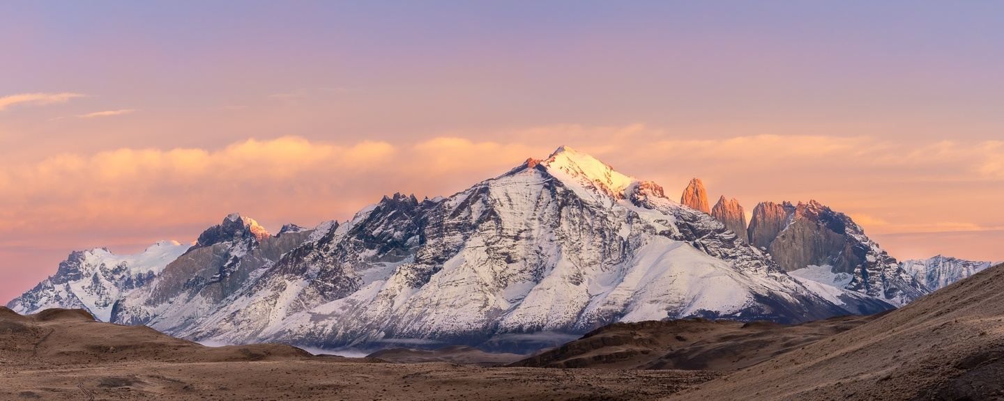 Snowy mountain range at sunset with pink-orange sky and dark foreground hills