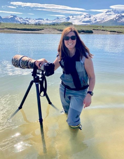 Woman wading in a mountain lake beside a tripod-mounted camera, with snowy peaks in the background