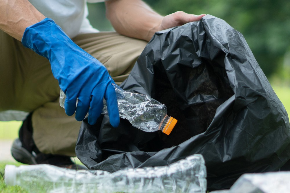 Gloved hands collecting trash into a black bag outdoors, with a plastic bottle in the foreground