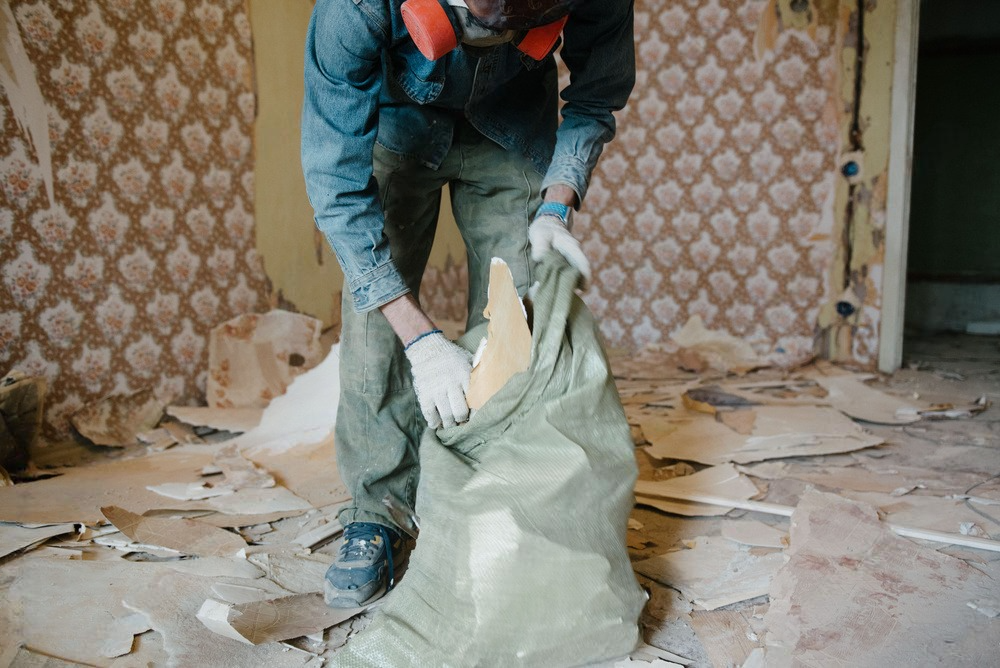 Worker in gloves empties debris into a green bag in a dusty room with torn wallpaper.