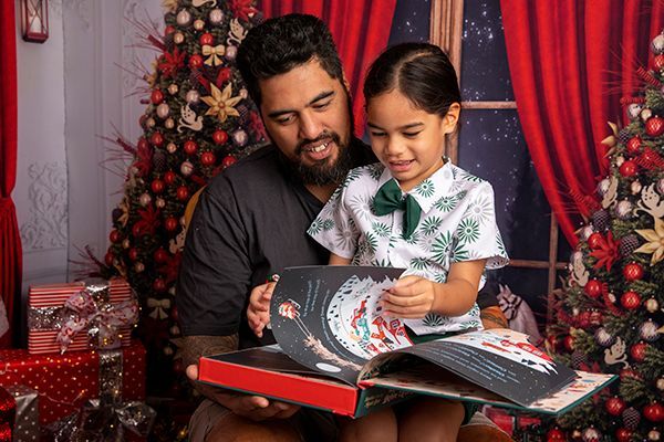 A man and a little girl are reading a book together in front of a christmas tree.