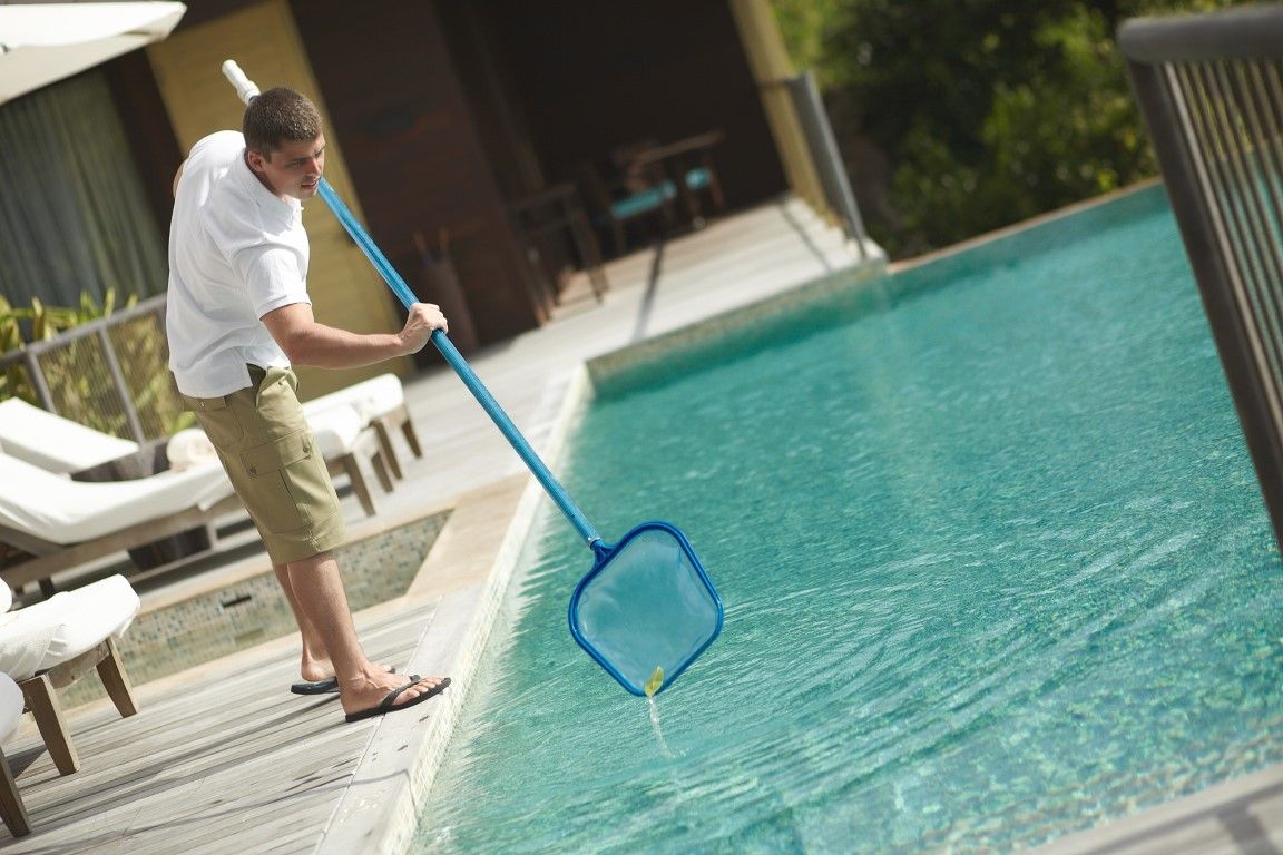 Person skimming a turquoise swimming pool with a blue net beside white lounge chairs