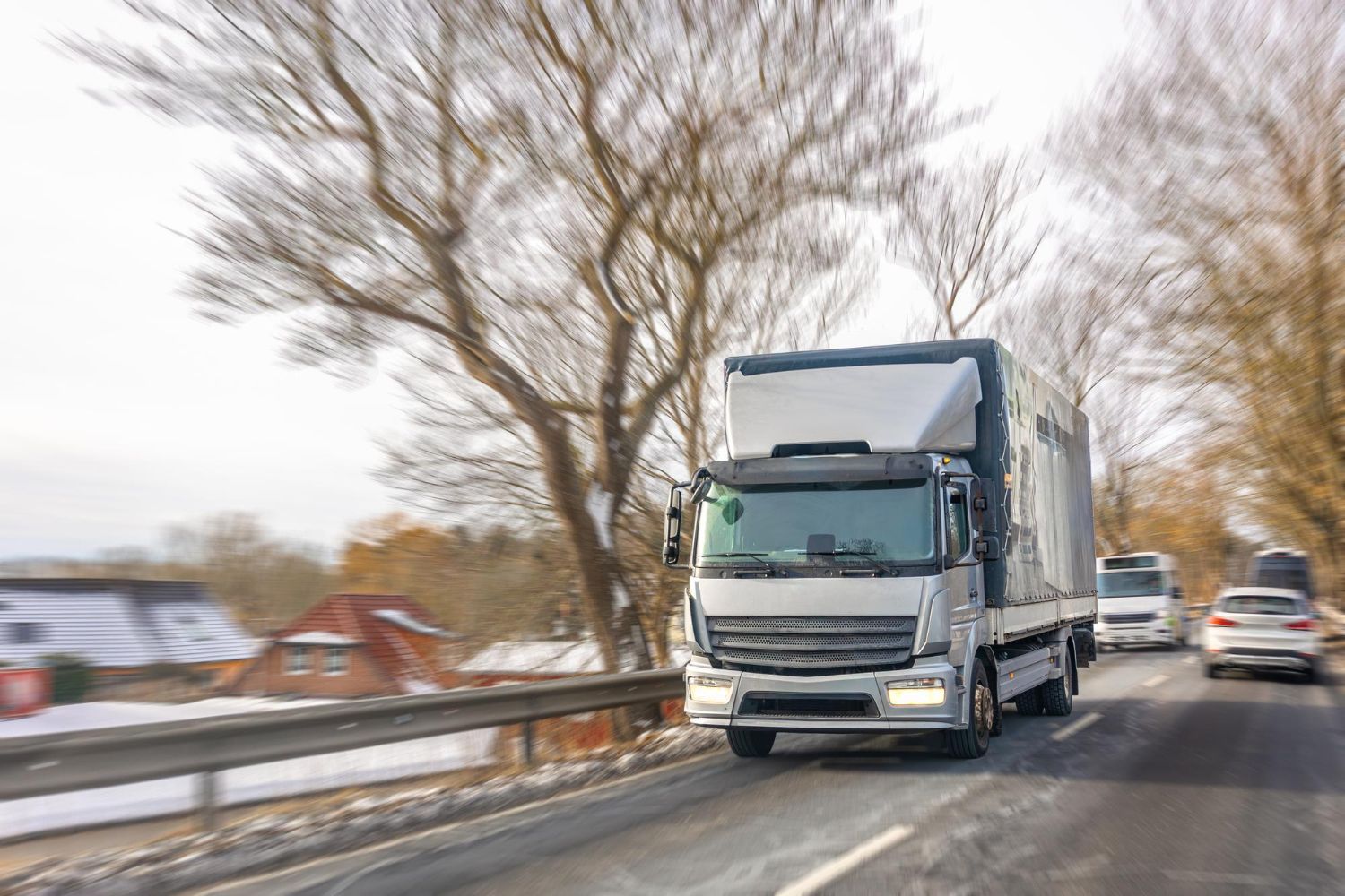White delivery truck driving on a snowy rural road with bare trees and cars behind it