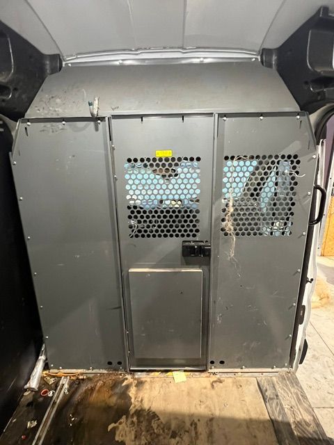 Inside a cargo van, a gray metal pet crate with mesh vents sits against the back doors.