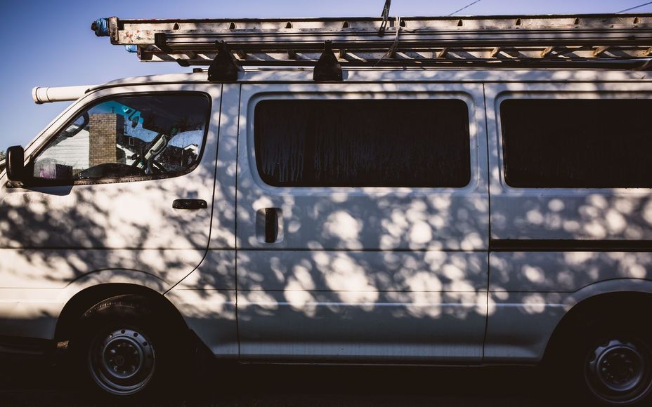 White cargo van parked on a street, with a roof rack and tree shadows across its side.