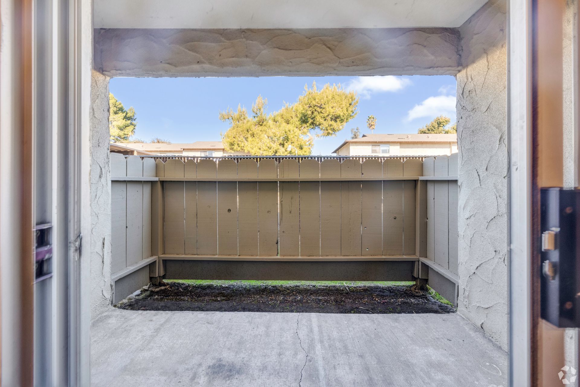 View from doorway to a small, walled patio. The sky is blue, and a tree is visible beyond the wall.