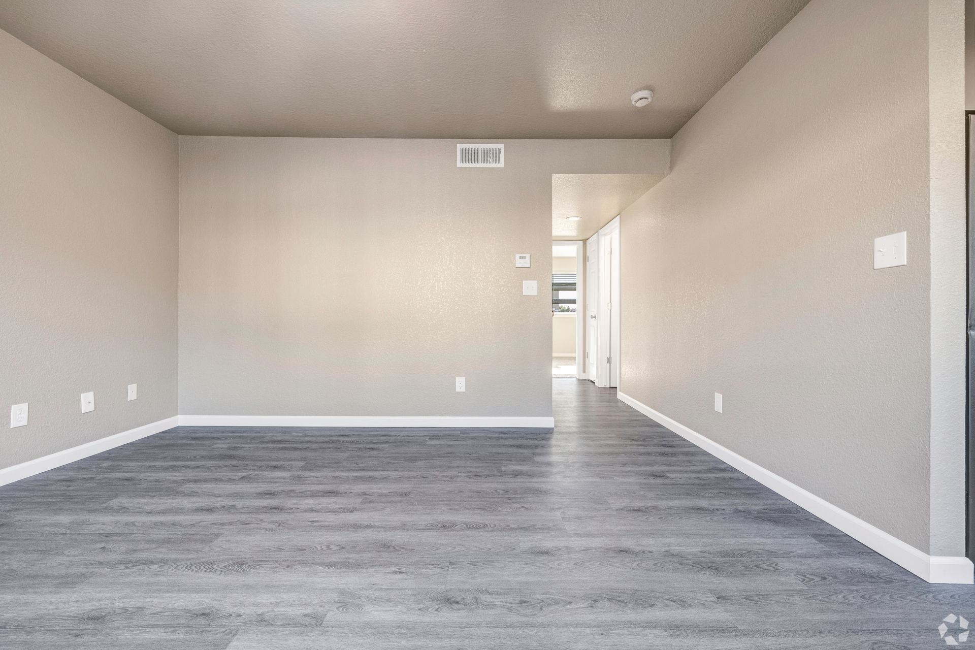 Empty living room with gray walls and wood-look flooring, leading to a hallway.